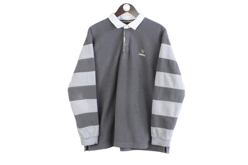 Puede incluir: Una camiseta de rugby gris con mangas largas a rayas blancas y grises. Tiene cuello blanco y un peque&ntilde;o logo en el pecho. La camiseta est&aacute; colgada de una percha de madera.