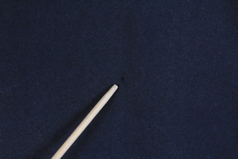 May include: A close-up of a dark navy blue fabric with a light-colored wooden stick diagonally across the frame. The fabric has a slightly textured appearance. The stick is smooth and cylindrical.