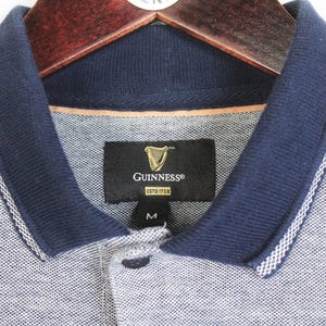 May include: A gray polo shirt with a navy blue collar and trim, hanging on a wooden hanger. The shirt features the Guinness logo on a black tag. The collar has a white stripe detail. The shirt is a size medium.