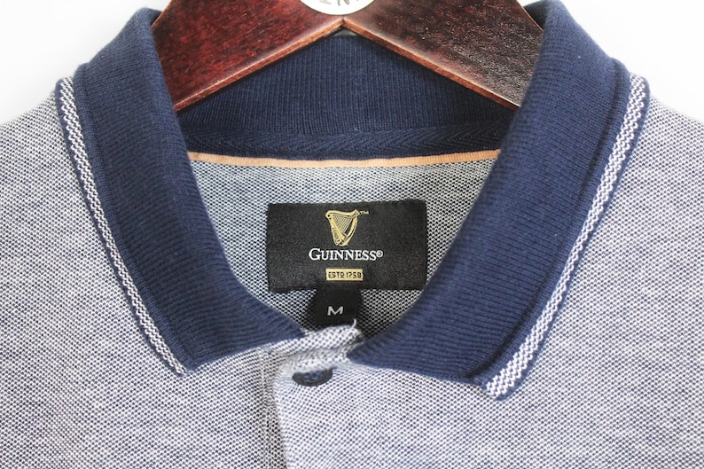 May include: A gray polo shirt with a navy blue collar and trim, hanging on a wooden hanger. The shirt features the Guinness logo on a black tag. The collar has a white stripe detail. The shirt is a size medium.