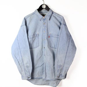 May include: A light blue long-sleeve denim shirt featuring a standard collar, a full-length button-down front, and two chest pockets. The shirt includes a small red tab detail on the left chest pocket and buttoned cuffs on the long sleeves.