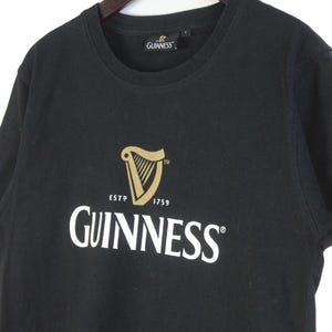 以下が含まれることがあります： ギネスのロゴが入った黒いTシャツ。ロゴには金のハープ、「ESTD 1759」の文字、白で「GUINNESS」の文字が入っています。シャツは柔らかい素材でできており、クルーネックです。