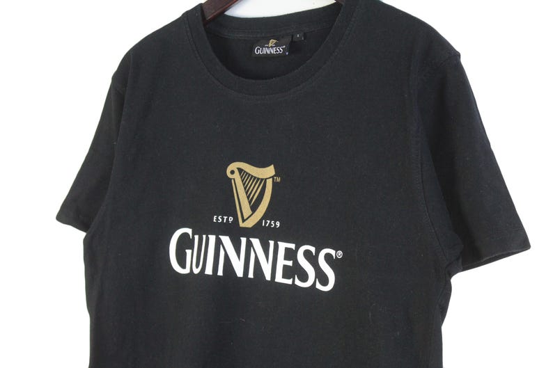 以下が含まれることがあります： ギネスのロゴが入った黒いTシャツ。ロゴには金のハープ、「ESTD 1759」の文字、白で「GUINNESS」の文字が入っています。シャツは柔らかい素材でできており、クルーネックです。
