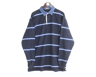 May include: Navy blue long-sleeve rugby shirt with horizontal white and light blue stripes. The shirt has a denim collar and a button-up placket. The shirt is hanging on a wooden hanger against a white background.