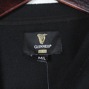 Op de afbeelding: Zwarte fleecejas met een Guinness-label met het logo van een gouden harp en de tekst "GUINNESS ESTP 1759". Het label geeft ook maat M/L aan. De jas hangt aan een houten kleerhanger.