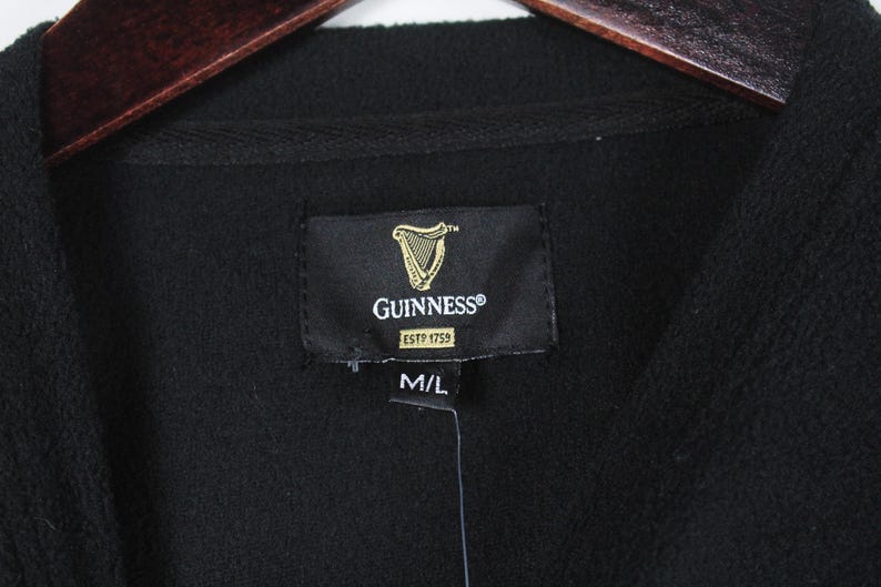 Op de afbeelding: Zwarte fleecejas met een Guinness-label met het logo van een gouden harp en de tekst "GUINNESS ESTP 1759". Het label geeft ook maat M/L aan. De jas hangt aan een houten kleerhanger.