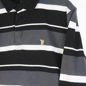 May include: A long-sleeved polo shirt with horizontal stripes in black, white, and gray. The shirt features a black collar and a small gold-colored emblem on the chest. The shirt is made of a soft material.