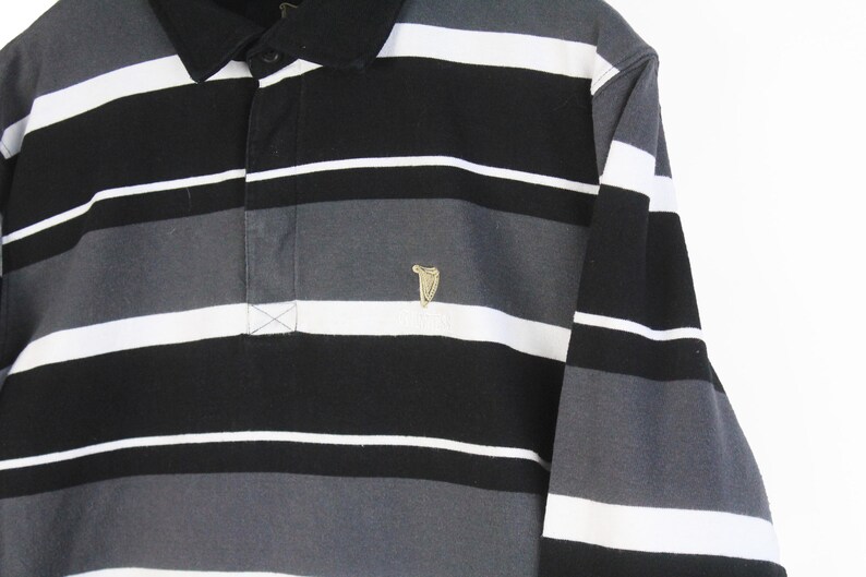 May include: A long-sleeved polo shirt with horizontal stripes in black, white, and gray. The shirt features a black collar and a small gold-colored emblem on the chest. The shirt is made of a soft material.