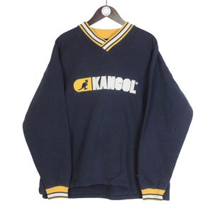 K&ouml;nnte beinhalten: Marineblaues Langarm-Sweatshirt mit V-Ausschnitt und dem KANGOL-Logo in Wei&szlig; und Gelb. Das Logo zeigt ein K&auml;nguru. Kragen und Manschetten haben gelbe und wei&szlig;e Streifen. Das Sweatshirt h&auml;ngt an einem Holzb&uuml;gel.