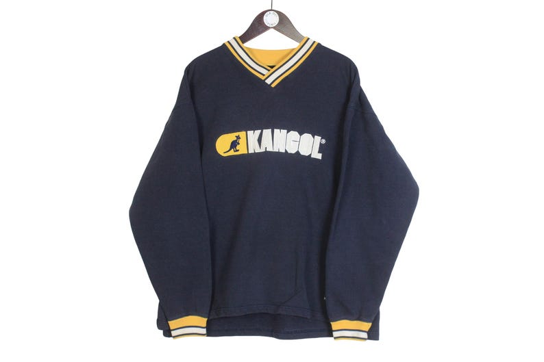 K&ouml;nnte beinhalten: Marineblaues Langarm-Sweatshirt mit V-Ausschnitt und dem KANGOL-Logo in Wei&szlig; und Gelb. Das Logo zeigt ein K&auml;nguru. Kragen und Manschetten haben gelbe und wei&szlig;e Streifen. Das Sweatshirt h&auml;ngt an einem Holzb&uuml;gel.