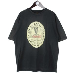 May include: Black t-shirt featuring a large oval graphic with the Guinness Extra Stout logo. The logo includes a harp and text that reads "St. James Gate Dublin". The shirt is hanging on a wooden hanger.