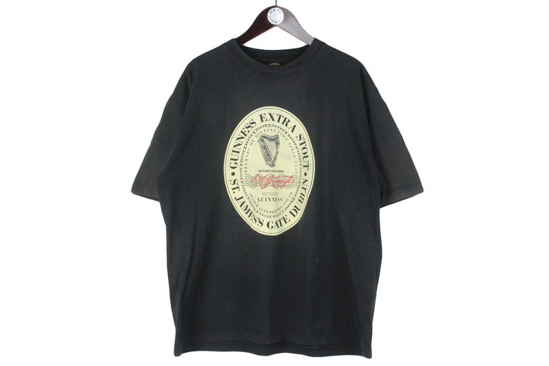 May include: Black t-shirt featuring a large oval graphic with the Guinness Extra Stout logo. The logo includes a harp and text that reads "St. James Gate Dublin". The shirt is hanging on a wooden hanger.