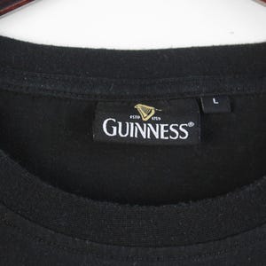 以下が含まれることがあります： クルーネックの黒いTシャツ。Guinnessのラベルには、ブランド名が白で表示され、ハープのロゴと「ESTD 1759」の文字が記されています。サイズラベルには「L」と記載されています。