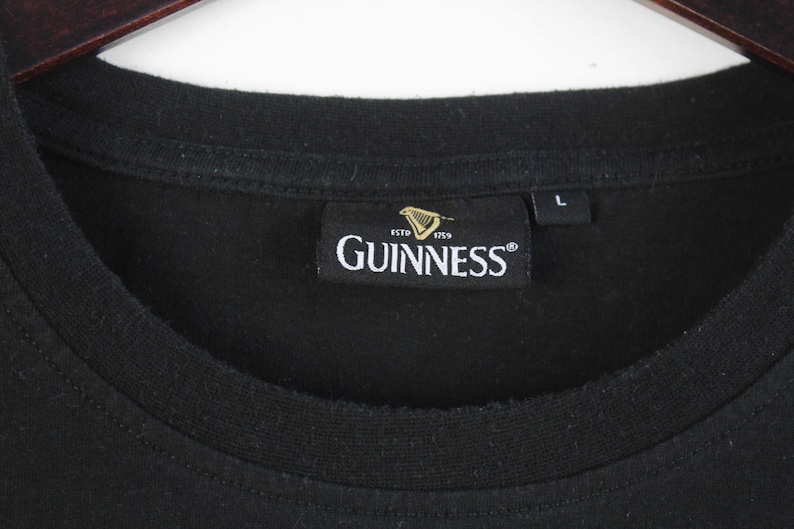 以下が含まれることがあります： クルーネックの黒いTシャツ。Guinnessのラベルには、ブランド名が白で表示され、ハープのロゴと「ESTD 1759」の文字が記されています。サイズラベルには「L」と記載されています。