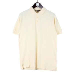 May include: Cream-coloured polo shirt with short sleeves, a collared neckline, and three buttons. The shirt is hanging on a black hanger against a white background. A classic, casual top.