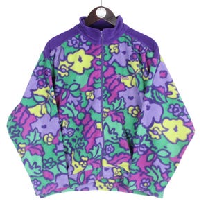 Puede incluir: Una chaqueta de forro polar de manga larga con cremallera completa y cuello alto. La chaqueta presenta un estampado floral multicolor vibrante en tonos morado, verde y amarillo. El logotipo de Fila est&aacute; bordado en el pecho izquierdo.