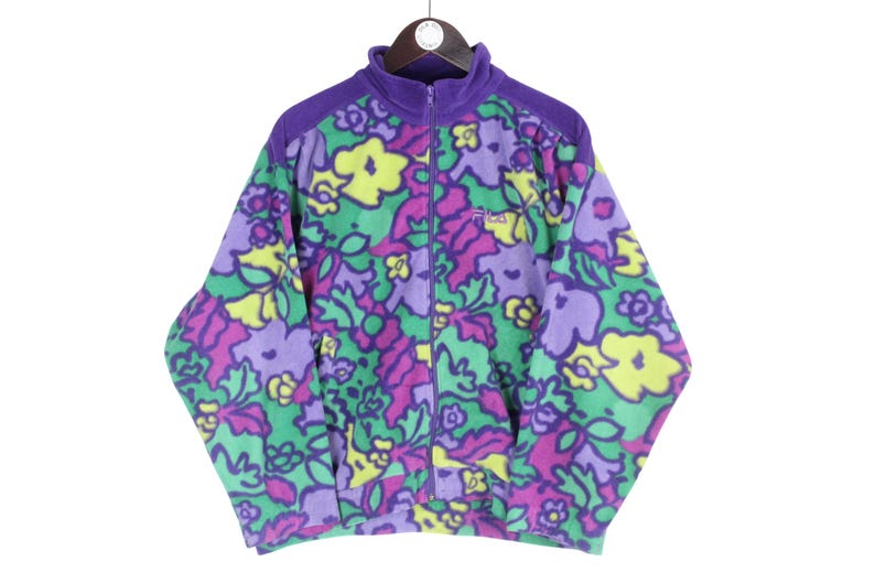 Puede incluir: Una chaqueta de forro polar de manga larga con cremallera completa y cuello alto. La chaqueta presenta un estampado floral multicolor vibrante en tonos morado, verde y amarillo. El logotipo de Fila est&aacute; bordado en el pecho izquierdo.