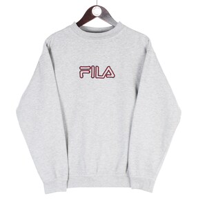 Puede incluir: Una sudadera gris claro con el nombre de la marca "FILA" en letras granates en el pecho. La sudadera est&aacute; colgada de una percha de madera sobre un fondo blanco. La parte superior de manga larga tiene cuello redondo.
