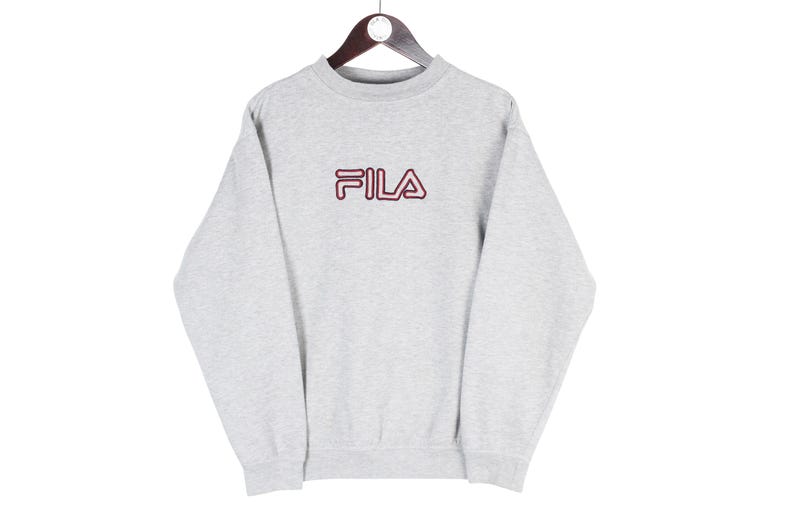 Puede incluir: Una sudadera gris claro con el nombre de la marca "FILA" en letras granates en el pecho. La sudadera est&aacute; colgada de una percha de madera sobre un fondo blanco. La parte superior de manga larga tiene cuello redondo.