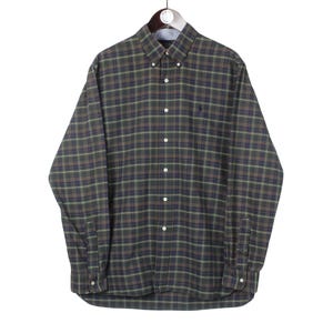 May include: A green, brown, and blue plaid long-sleeved shirt with a button-down collar. The shirt features a small embroidered logo on the chest. It is displayed on a hanger against a white backdrop.