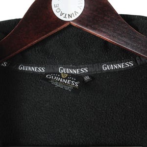 K&ouml;nnte beinhalten: Schwarze Fleecejacke mit Rei&szlig;verschluss und Guinness-Logo auf der Vorderseite. Die Jacke hat ein schwarzes Etikett mit dem Guinness-Logo und dem Text "Est. 1778 Guinness Merchandise" darauf.