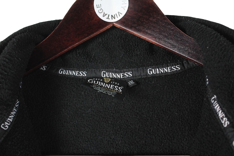 K&ouml;nnte beinhalten: Schwarze Fleecejacke mit Rei&szlig;verschluss und Guinness-Logo auf der Vorderseite. Die Jacke hat ein schwarzes Etikett mit dem Guinness-Logo und dem Text "Est. 1778 Guinness Merchandise" darauf.