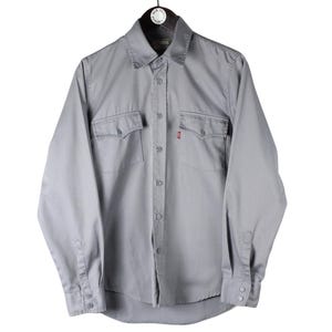 May include: A light grey long-sleeve button-down shirt featuring a standard collar, a front button placket, and two chest pockets with buttoned flaps. A small red tab is visible on the edge of the left chest pocket. The shirt has long sleeves.