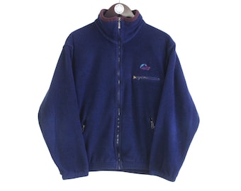 May include: A navy blue fleece jacket with a full-length zipper and a small chest pocket. The jacket has a high collar with a burgundy lining and a small logo on the chest. The jacket is hanging on a hanger.