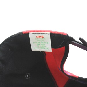 May include: Black Nike baseball cap with red, green, and pink accents. The cap features a curved brim and an adjustable strap. A white tag with the Nike logo and product information is visible.