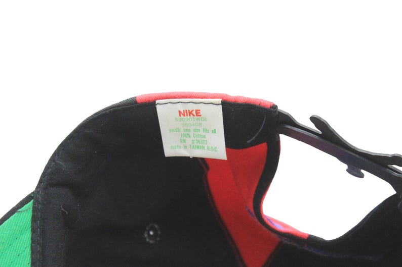 May include: Black Nike baseball cap with red, green, and pink accents. The cap features a curved brim and an adjustable strap. A white tag with the Nike logo and product information is visible.