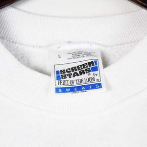 May include: Close-up of a white sweatshirt collar with a tag. The tag is rectangular with blue and white text that reads "SCREEN STARS by FRUIT OF THE LOOM SWEATS STYLED IN USA". The sweatshirt is a size "L".