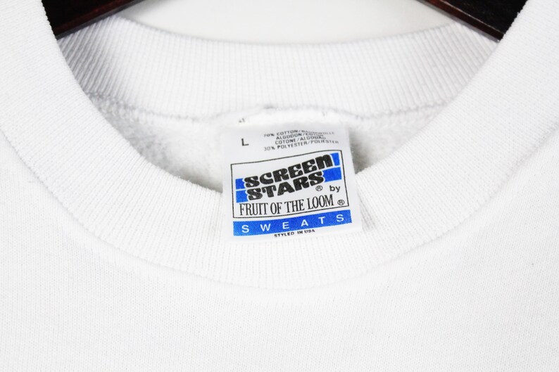 May include: Close-up of a white sweatshirt collar with a tag. The tag is rectangular with blue and white text that reads "SCREEN STARS by FRUIT OF THE LOOM SWEATS STYLED IN USA". The sweatshirt is a size "L".