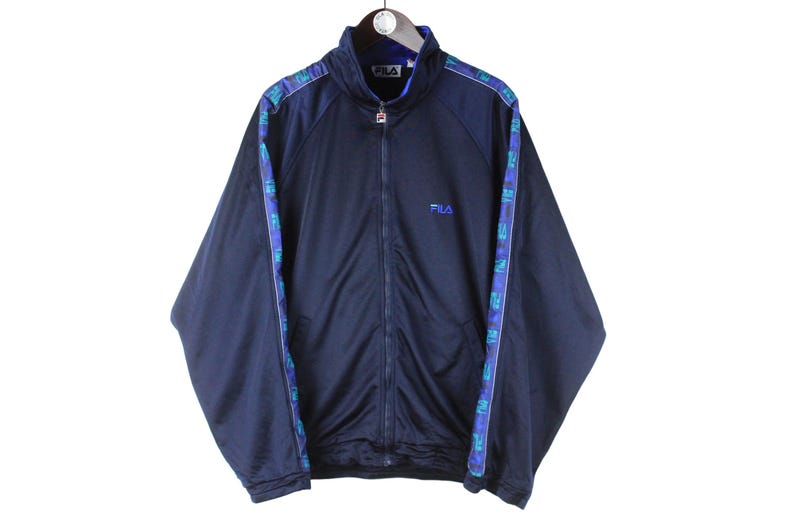 Puede incluir: Una chaqueta azul marino con cremallera con un ribete estampado en azul y verde. La chaqueta tiene un logotipo de Fila en el pecho izquierdo.