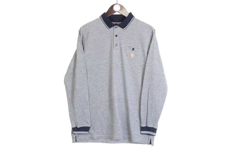 May include: A long-sleeved gray polo shirt with a navy blue collar, cuffs, and sleeve trim. The shirt has a three-button placket and a small pocket on the chest with a gold emblem. The shirt is hanging on a wooden hanger.