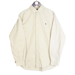 Può includere: Camicia beige chiaro a maniche lunghe, abbottonata e appesa a una gruccia di legno. La camicia ha un colletto classico, polsini abbottonati e un piccolo logo blu sul petto. La camicia è realizzata in tessuto liscio.