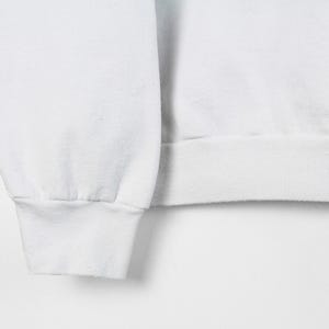 May include: Close-up of a white sweatshirt sleeve and cuff. The cuff is ribbed and the sleeve is smooth. The fabric appears to be a soft, medium-weight material. The background is plain white.