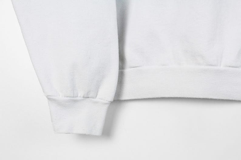 May include: Close-up of a white sweatshirt sleeve and cuff. The cuff is ribbed and the sleeve is smooth. The fabric appears to be a soft, medium-weight material. The background is plain white.