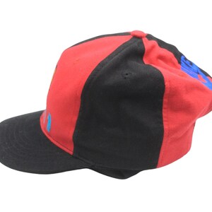 May include: A red and black baseball cap with a curved brim. The cap features a color block design with red on the top and black on the sides and brim. Blue text is visible on the side of the cap.
