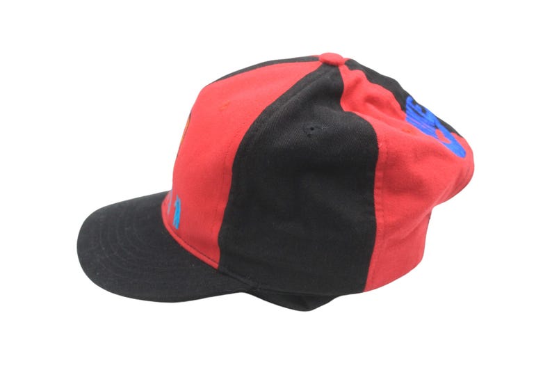 May include: A red and black baseball cap with a curved brim. The cap features a color block design with red on the top and black on the sides and brim. Blue text is visible on the side of the cap.
