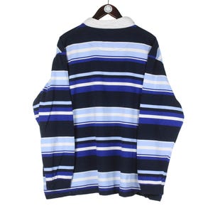 Puede incluir: Una camiseta de rugby de manga larga con rayas horizontales en azul marino, azul claro y blanco. La camiseta tiene un cuello blanco y est&aacute; colgada de una percha de madera. La parte trasera de la camiseta es visible.