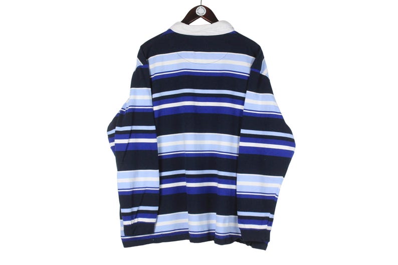 Puede incluir: Una camiseta de rugby de manga larga con rayas horizontales en azul marino, azul claro y blanco. La camiseta tiene un cuello blanco y est&aacute; colgada de una percha de madera. La parte trasera de la camiseta es visible.