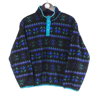 May include: Black fleece pullover with a geometric pattern in teal, purple, and blue. Features a teal collar and button placket. The pullover is hanging on a black hanger.