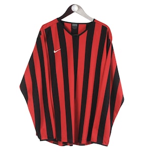 Puede incluir: Camiseta de manga larga a rayas rojas y negras colgada de una percha. La camiseta tiene cuello en V y un peque&ntilde;o logotipo blanco de Nike en el pecho. La camiseta est&aacute; hecha de un material ligero.