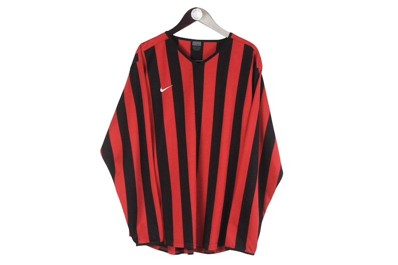 Puede incluir: Camiseta de manga larga a rayas rojas y negras colgada de una percha. La camiseta tiene cuello en V y un peque&ntilde;o logotipo blanco de Nike en el pecho. La camiseta est&aacute; hecha de un material ligero.