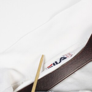 May include: Close-up of a white garment with a Fila logo tag. The tag features the Fila logo in red, navy, and white. The garment is hanging on a wooden hanger with a DLA Vintage tag.