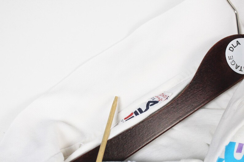May include: Close-up of a white garment with a Fila logo tag. The tag features the Fila logo in red, navy, and white. The garment is hanging on a wooden hanger with a DLA Vintage tag.