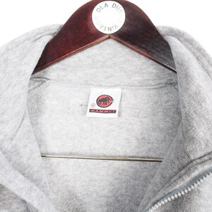 Pu&ograve; includere: Primo piano di una giacca in pile grigia con cerniera intera e collo alto. La giacca presenta una toppa con il logo Mammut sul petto. &Egrave; appesa a una gruccia di legno.