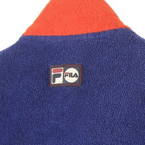 May include: Close-up of a blue fleece jacket with an orange collar. The jacket features a Fila logo patch on the upper back. The fleece material has a textured appearance.