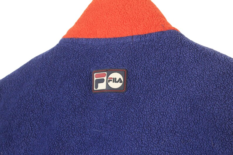 May include: Close-up of a blue fleece jacket with an orange collar. The jacket features a Fila logo patch on the upper back. The fleece material has a textured appearance.