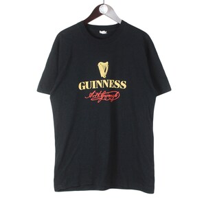 Puede incluir: Camiseta negra con el logo de Guinness, que incluye un arpa dorada y texto. La palabra "Guinness" es dorada, con la firma en rojo. La camiseta est&aacute; colgada en una percha de madera.
